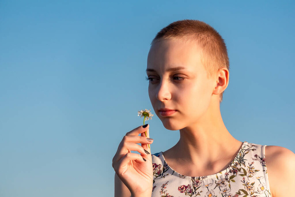 portrait of a teenage girl with a short haircut in a dress holding a camomile in her hand against the sk - Foto, Imagen