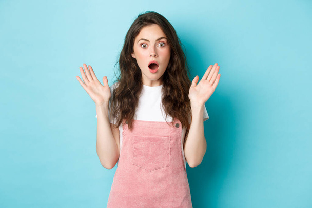 Portrait of shocked attractive woman gasping, spread hands sideways and stare startled at camera, hear worrying terrible news, looking with disbelief, standing against bue background - Foto, Bild