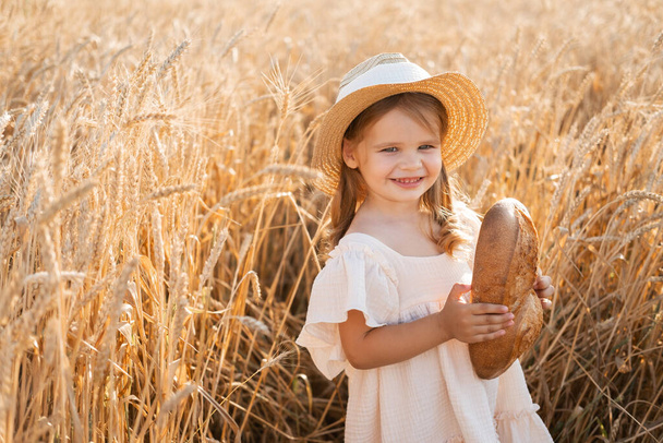 little blonde girl in straw hat and beige muslin dress holds bread in wheat field on sunset - Photo, image