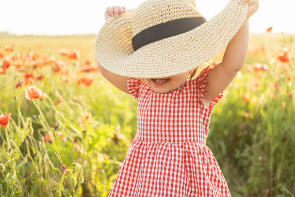 χαριτωμένο κοριτσάκι με κόκκινο φόρεμα και ψάθινο καπέλο στον τομέα της παπαρούνας στο ηλιοβασίλεμα του καλοκαιριού - Φωτογραφία, εικόνα