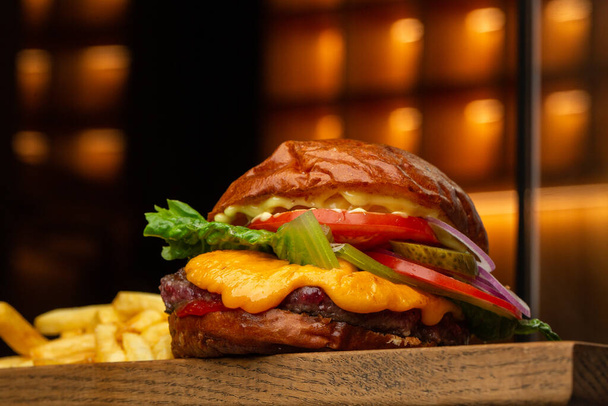 Big cheeseburger with french fries isolated on background with yellow light. Hamburger with beef patty onion, tomato, lettuce pickles, melted cheddar, mustards, tomato jam, mayo. - Foto, imagen