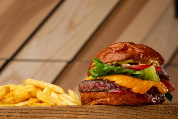 Big cheeseburger with french fries isolated on wooden background. Hamburger with beef patty onion, tomato, lettuce pickles, melted cheddar, mustards, tomato jam, mayo. Horizontal - Foto, imagen