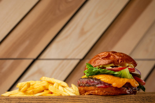 Big cheeseburger with french fries isolated on wooden background. Hamburger with beef patty onion, tomato, lettuce pickles, melted cheddar, mustards, tomato jam, mayo. Horizontal - Fotografie, Obrázek