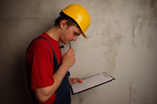 Der Arbeiter untersucht sorgfältig den Plan der Wohnung. Junger Bauarbeiter in rotem T-Shirt, blauem Gewand und gelbem Bauhelm auf dem Hintergrund einer Betonwand. Sanierungskonzept. - Foto, Bild