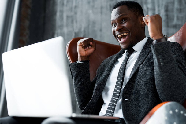 Cheerful african american businessman sitting on a chair and working at a laptop rejoicing - Фото, зображення