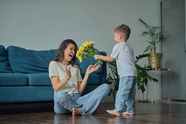 Ευτυχισμένη ημέρα της μητέρας. Παιδί γιος συγχαίρει τη μητέρα για τις διακοπές και δίνει λουλούδια. συγχαίροντάς την για την ημέρα των μητέρων κατά τη διάρκεια των διακοπών στο σπίτι - Φωτογραφία, εικόνα