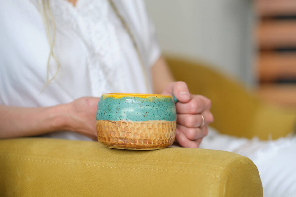 unrecognizable woman in white clothes sits on a yellow sofa and holds a yellow handmade cup. - Photo, image