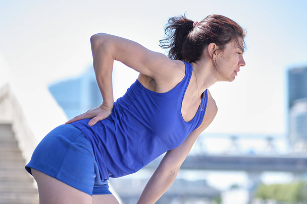 Athletic woman in blue sportswear rubbing the muscles of her lower back, portrait. Back pain concept. - 写真・画像