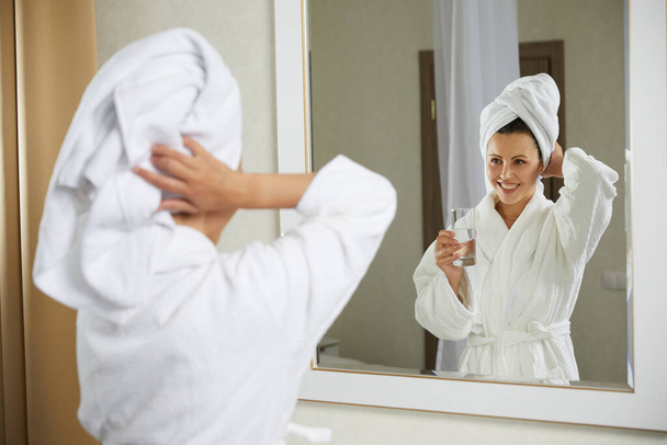 Beautiful happy girl drinking and holding glass of water standing in front of mirror in the morning. Girl wearing robe after taking shower - Zdjęcie, obraz