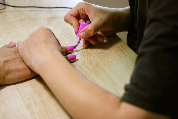 Ένας μανικιουρίστας εφαρμόζει ροζ βερνίκι νυχιών με ένα πινέλο στα νύχια ενός πελάτη σε ένα σαλόνι ομορφιάς. Θεραπείες σπα. Γυναικεία χέρια και δάχτυλα στο ξύλινο τραπέζι από κοντά. Γυναικεία διαδικασία φροντίδας νυχιών.  - Φωτογραφία, εικόνα