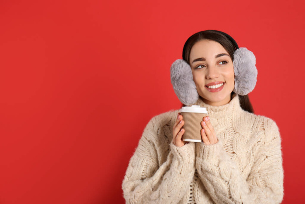 Beautiful young woman in earmuffs with cup of drink on red background. Space for text - Photo, Image