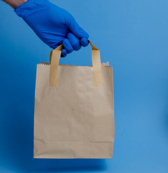 brown paper shopping bag on blue background. Brown empty craft lunch bag. Recycle brown paper bag. Hand in glove holding a paper bag.  - Zdjęcie, obraz