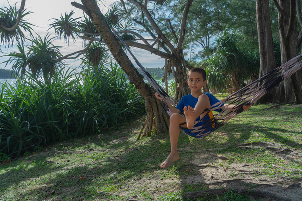 Poika sinisessä mekossa rentouttava riippumatossa varjossa mänty meren rannalla - Valokuva, kuva