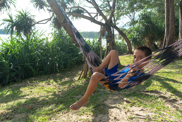 Mavi elbiseli bir çocuk deniz kenarındaki çam ağacının gölgesinde hamakta dinleniyor. - Fotoğraf, Görsel