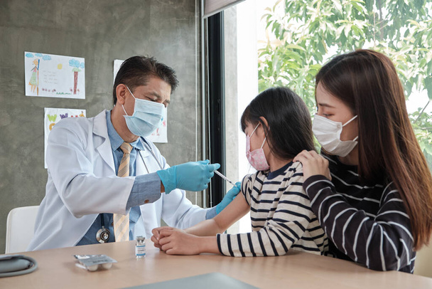 Чоловік-лікар з маскою обличчя вакцинований азіатською дівчиною, щоб запобігти коронавірусу (COVID) 19) в педіатричній клініці в дитячій лікарні з матір'ю поруч. Інфекції лікують хвороби, завдають дітям болю.. - Фото, зображення