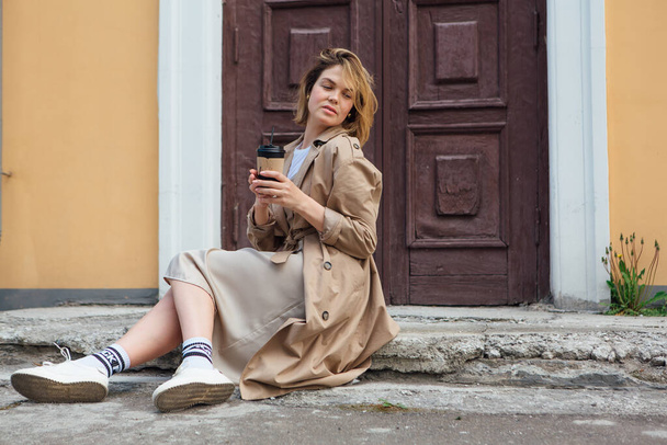 Young smiling millennial woman with wild hair dressed in an autumn coat sitting near the door of an old building with a cup of coffee to go. - Photo, image