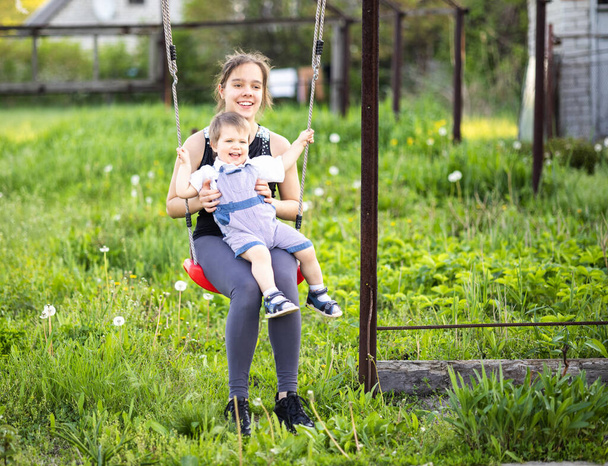 Cute brother and older sister ride on a bright swing on warm spring day against the backdrop of a green blooming garden - Фото, зображення