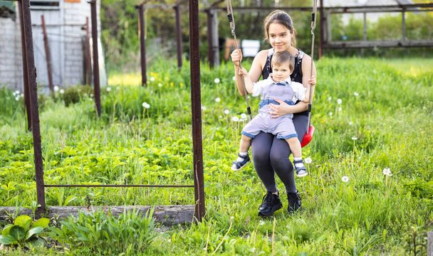 Cute brother and older sister ride on a bright swing warm spring day against the backdrop of a green blooming garden - Foto, imagen