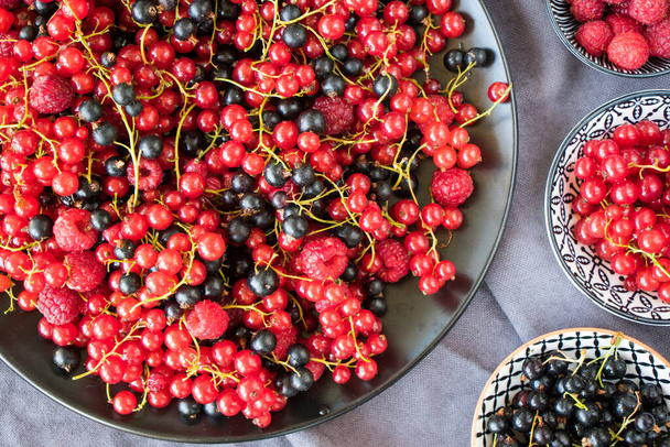 Red and black currant and loganberries on the black plate and gray cloth background. Large group of colorful berries. - Photo, Image
