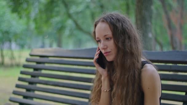 kaupallinen ammunta, nuori tyttö puistossa istuu penkillä mustassa mekossa ja puhuu puhelimessa. - Materiaali, video