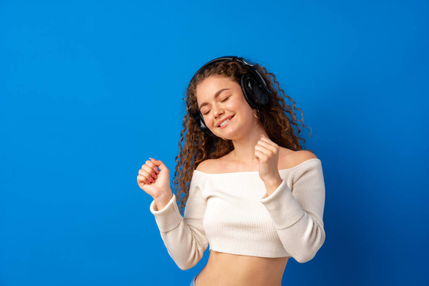 Young attractive curly woman listening to music with headphones against blue background - Φωτογραφία, εικόνα