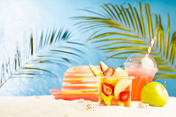 Resort lifestyle - picnic with cold cocktail, fresh tropical fruit salad, sun hat on white sand with palm, sun glare in blue sea view. Vacation on ocean beach.  - Foto, imagen