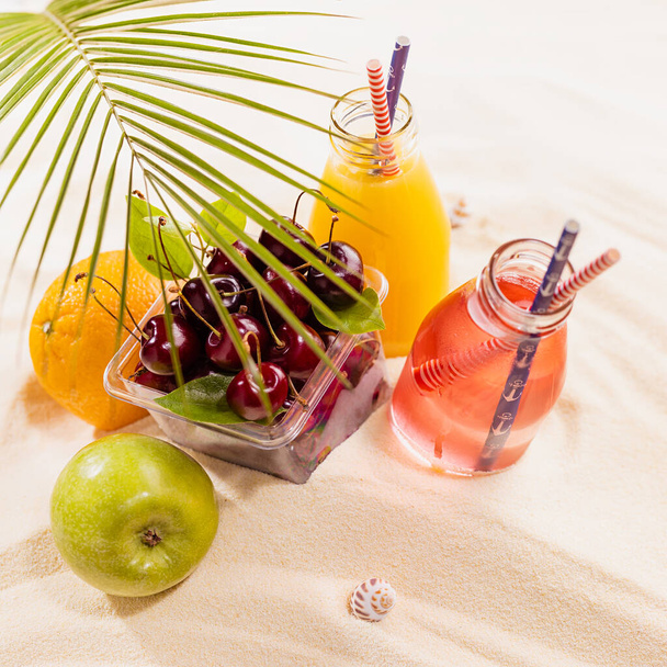 Summer lifestyle background picnic with drink and fresh fruit on ocean coast - cold juicy beverages, green palm leaf, red cherries, orange, apple on white sand, square. - Foto, imagen