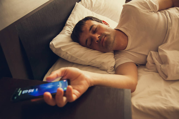 Een man wordt wakker in bed en kijkt naar de telefoon, zet het alarm uit en kijkt naar de tijd, surft op het internet, leest berichten, controleert sociale netwerken. - Foto, afbeelding