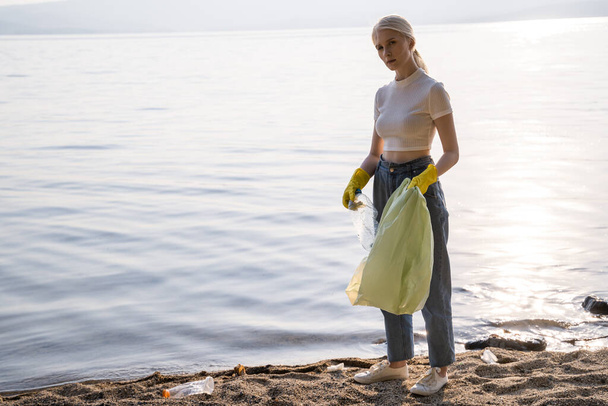 A woman in gloves stands on the shore of the lake and looks into the camera with annoyance. There is a lot of rubbish around it. - Foto, imagen