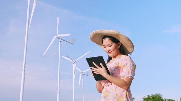 背景風力発電タービンのフィールドにデジタルタブレットを使用してアジアの若い女性. - 写真・画像