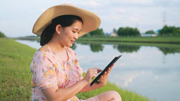 Mulher asiática relaxante com o uso de dispositivo portátil sentado no gramado perto do rio. - Foto, Imagem