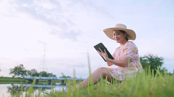 Mulher asiática relaxante com o uso de dispositivo portátil sentado no gramado perto do rio. - Foto, Imagem