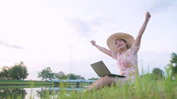 Hermosa mujer asiática en sombrero sentado en el césped trabajando en la computadora sensación de éxito. - Foto, imagen