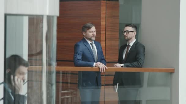 Medium slowmo shot of two confident male lawyers in suits having conversation in office while their colleague going down in glass elevator talking on phone - Footage, Video