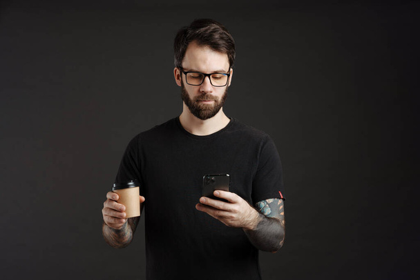 Bearded focused man using cellphone while drinking coffee takeaway isolated over black background - Foto, Imagen