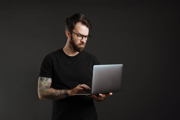 Bearded brunette man in eyeglasses think and using laptop isolated over black background - Zdjęcie, obraz