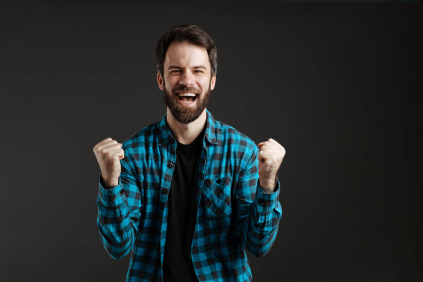 Bearded excite man in shirt laughing while making winner gesture isolated over black background - 写真・画像