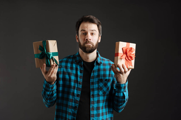 Bearded puzzled man looking at camera while showing gift boxes isolated over black background - Φωτογραφία, εικόνα