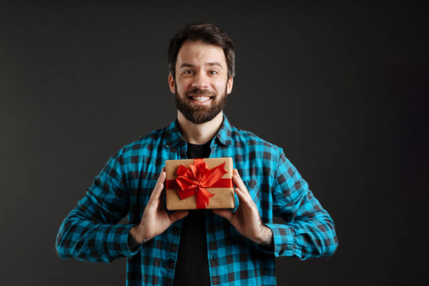 Bearded happy white man smiling while posing with gift box isolated over black background - Photo, image