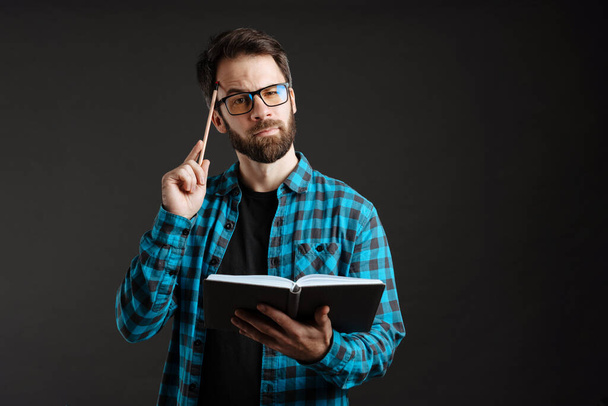 Bearded thinking man in eyeglasses posing with pencil and planner isolated over black background - Φωτογραφία, εικόνα