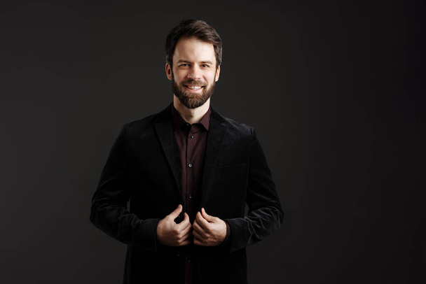Bearded happy man in black jacket smiling and looking at camera isolated over black background - Foto, afbeelding