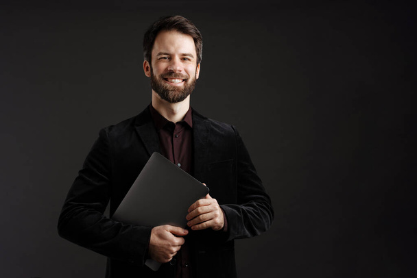 Bearded happy man in black jacket smiling while posing with laptop isolated over black background - Photo, image
