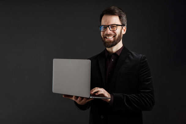 Bearded happy man in jacket smiling while using laptop isolated over black background - Zdjęcie, obraz
