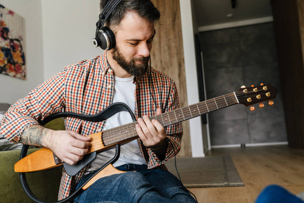 Smiling mid aged brunette bearded man playing acoustic guitar while sitting on a floor in the living room, wearing headphones - Valokuva, kuva
