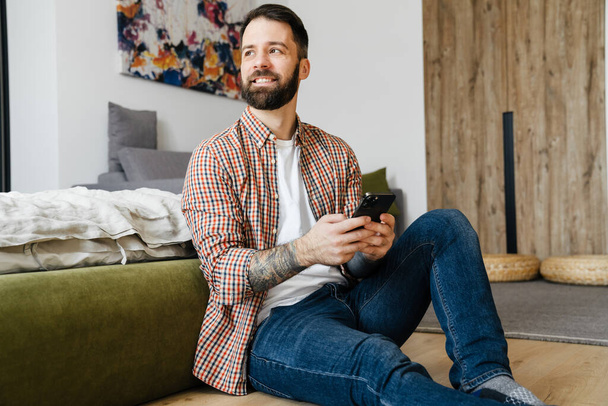 Smiling mid aged brunette bearded man holding mobile phone, sitting on a floor in the living room - Photo, image