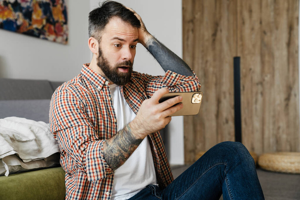Shocked mid aged brunette bearded man holding mobile phone, sitting on a floor in the living room - Photo, image
