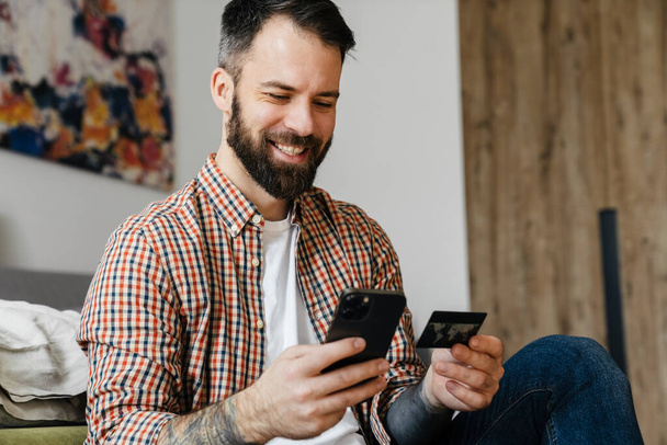 Smiling mid aged brunette bearded man holding mobile phone, sitting on a floor in the living room - Foto, afbeelding