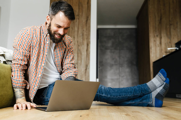 Happy mid aged brunette bearded man working on laptop computer while sitting on a floor in the living room - Photo, Image
