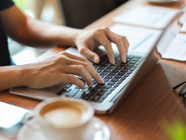 Mujer trabajadora escribiendo en el teclado del ordenador portátil con informe financiero y taza de café al lado en el escritorio - Foto, Imagen