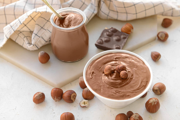 Jar and bowl with tasty chocolate paste and hazelnuts on light background - Photo, Image